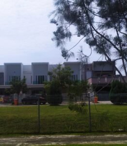 马来西亚住宅区内的两层排屋，前方有铁栅栏围篱、草坪及停放的车辆，背景为树木与灰白色天空